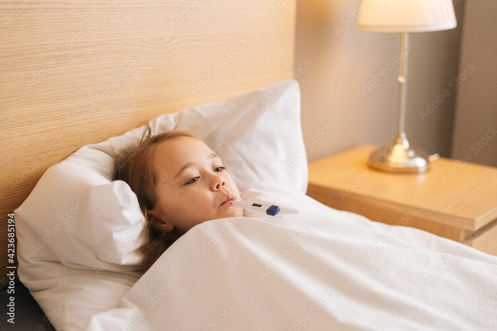 Close-up face of little sick girl lying in bed measuring temperature with thermometer in mouth at home, looking away. Unhappy child with high fever lying at bad in medical clinic. Close-up face of little sick girl lying in bed measuring temperature with thermometer in mouth at home, looking away. Unhappy child with high fever lying at bad in medical clinic.
