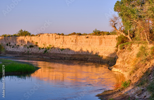 Calm river flow with delightful ripples in the water and golden reflection of clay cliffs against a blue sky at sunrise