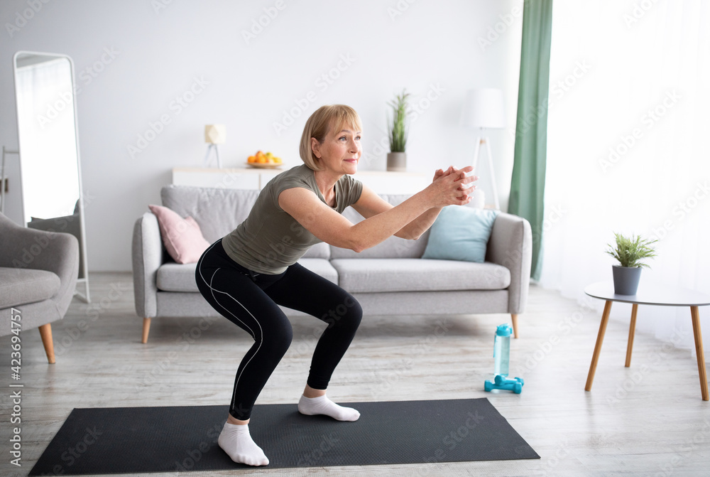 Positive senior lady doing squats on domestic workout in living room, free space Positive senior lady doing squats on domestic workout in living room, free space