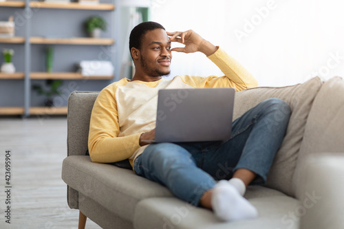 Dreamy black guy with laptop resting on sofa at home