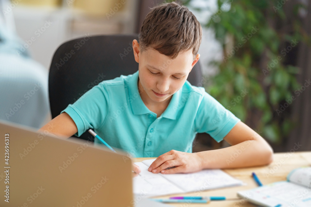 inquisitive boy uses laptop to study school subjects during his online lesson at home. Writing down topic lesson in notebook. E-Education Distance Home Education. inquisitive boy uses laptop to study school subjects during his online lesson at home. Writing down topic lesson in notebook. E-Education Distance Home Education.