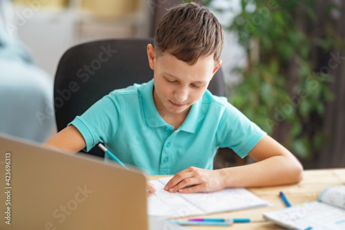 inquisitive boy uses laptop to study school subjects during his online lesson at home. Writing down topic lesson in notebook. E-Education Distance Home Education.