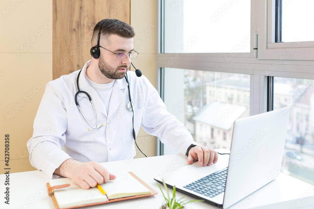 Man professional doctor in white medical coat and headset making conference call on laptop computer, consulting distance patient online in video chat, male explain treatment by webcam. Telemedicine Man professional doctor in white medical coat and headset making conference call on laptop computer, consulting distance patient online in video chat, male explain treatment by webcam. Telemedicine
