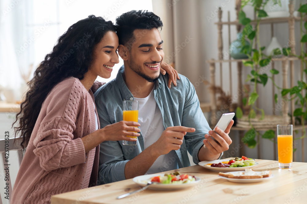 Cheerful Arab Spouses Using Smartphone While Eating Breakfast In Kitchen Cheerful Arab Spouses Using Smartphone While Eating Breakfast In Kitchen