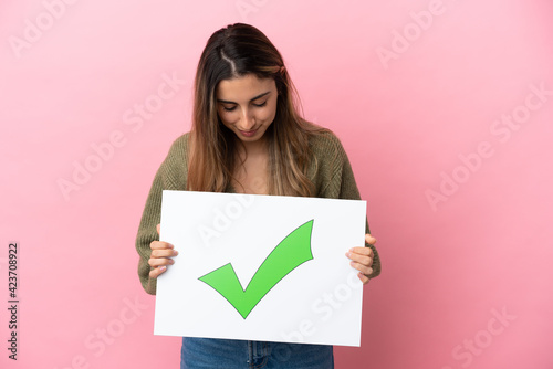 Young caucasian woman isolated on pink background holding a placard with text Green check mark icon