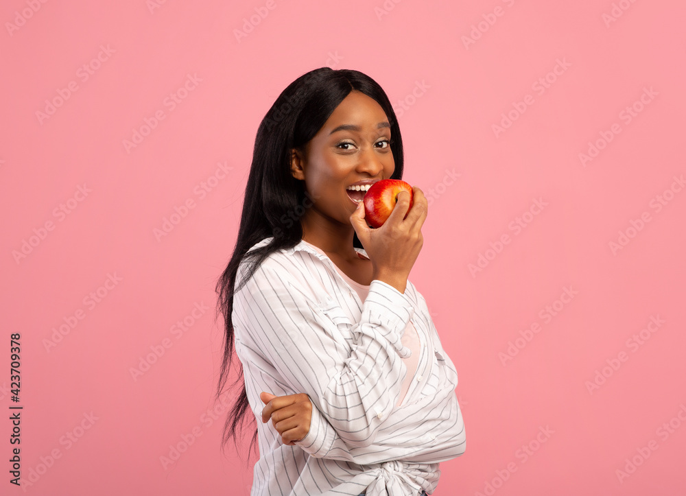 Positive black woman eating tasty red apple on pink studio background. Healthy lifestyle concept Positive black woman eating tasty red apple on pink studio background. Healthy lifestyle concept