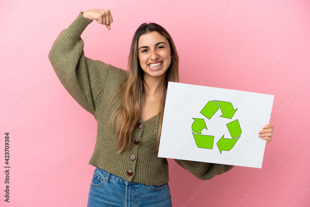 Young caucasian woman isolated on pink background holding a placard with recycle icon and doing strong gesture