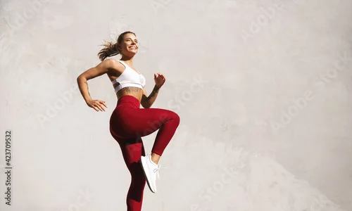 Obraz Fit woman exercising outdoors. Healthy young female athlete doing fitness workout. Sportswoman raising leg, do functional training outside on bright sunny day, smiling pleased, wearing sport outfit