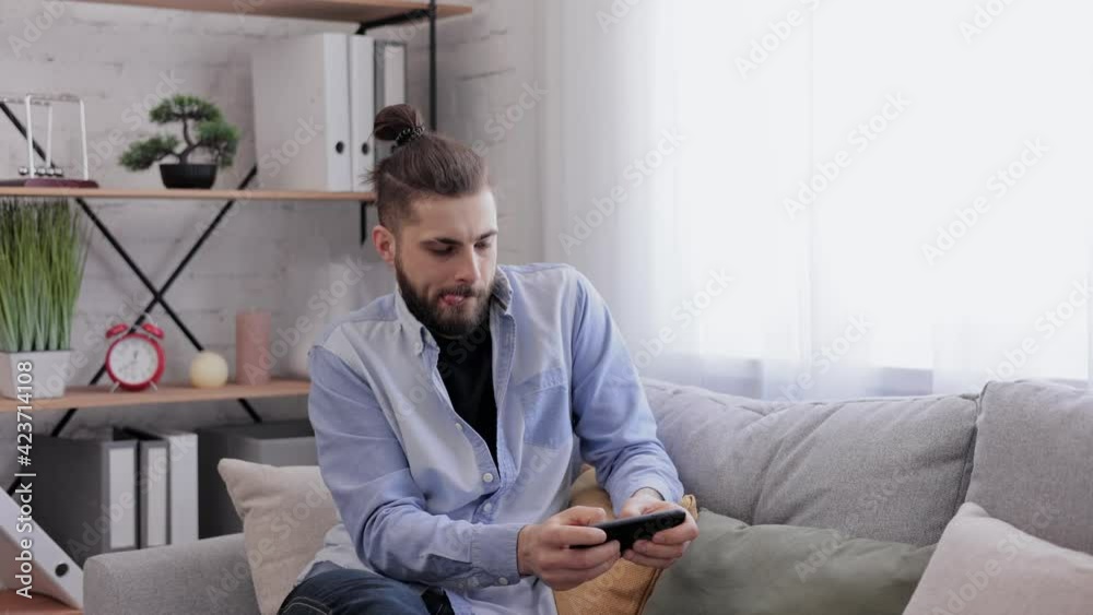 Cheerful young man playing video game on smartphone sitting on sofa at home. Handsome emotional guy celebrating victory in car racing game on mobile phone. Spending time at home, leisure, relax. Cheerful young man playing video game on smartphone sitting on sofa at home. Handsome emotional guy celebrating victory in car racing game on mobile phone. Spending time at home, leisure, relax.