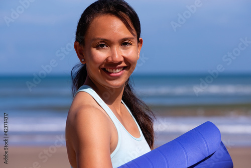 people, fitness, sport and healthy lifestyle concept - portrait of beautiful asian girl in sport clother with yoga mat on tropical beach, looking to the camera and smiling