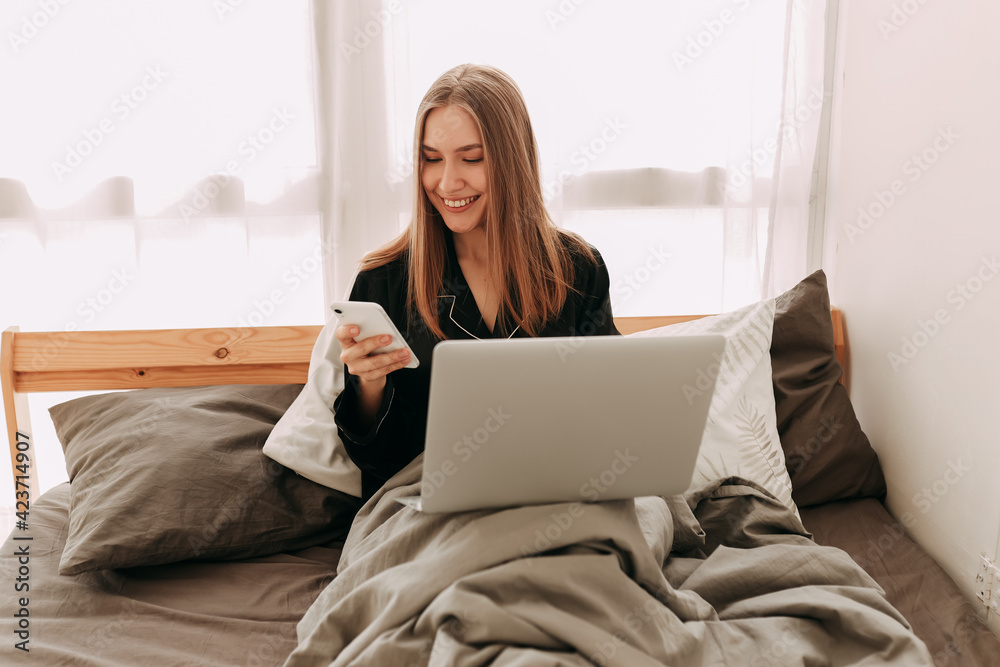 Smiling young business woman freelance student in domestic pajamas working remotely using technology and talking on a mobile phone looking at a laptop sitting in bed at home, selective focus Smiling young business woman freelance student in domestic pajamas working remotely using technology and talking on a mobile phone looking at a laptop sitting in bed at home, selective focus