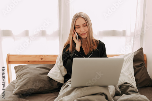Smiling young business woman freelance student in domestic pajamas working remotely using technology and talking on a mobile phone looking at a laptop sitting in bed at home, selective focus