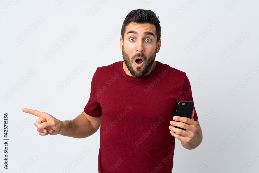 Young handsome man with beard using mobile phone isolated on white background surprised and pointing side Young handsome man with beard using mobile phone isolated on white background surprised and pointing side
