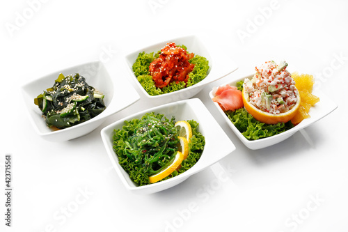Composition of oriental appetizers in bowls, isolated on a white background. A pickled seagrass Japanese salad, a wakame and cucumber salad with sesame seeds, a Korean kimchi and a crab sticks salad.
