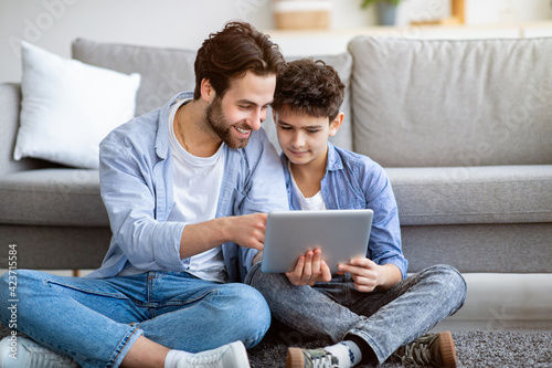 Father and son using tablet computer at home, checking new application, sitting on floor carpet near sofa