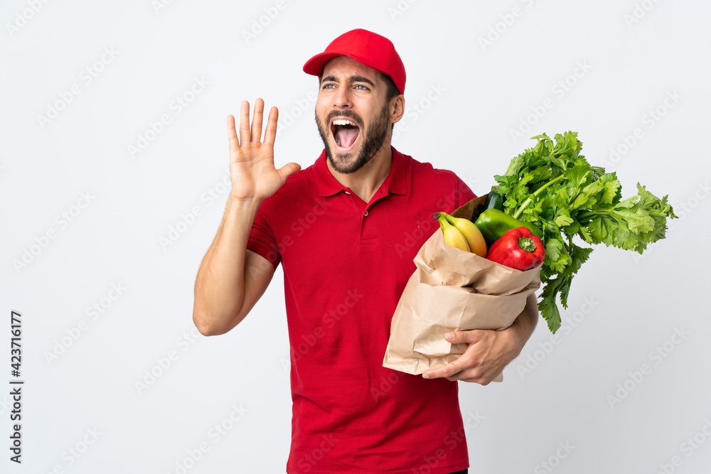 Young man with beard holding a bag full of vegetables isolated on white background shouting with mouth wide open