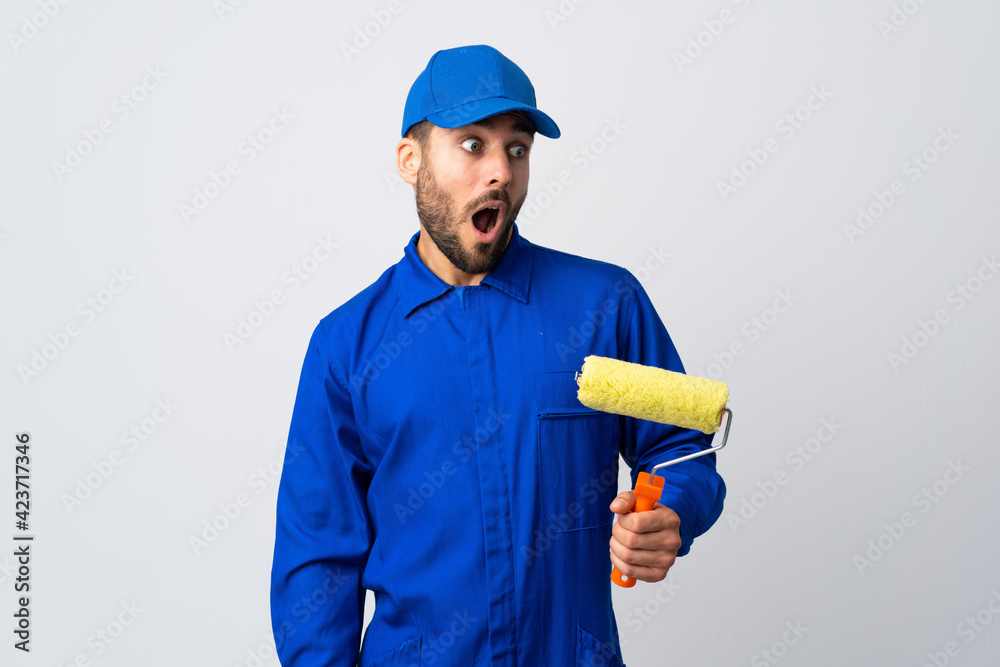 Painter man holding a paint roller isolated on white background doing surprise gesture while looking to the side