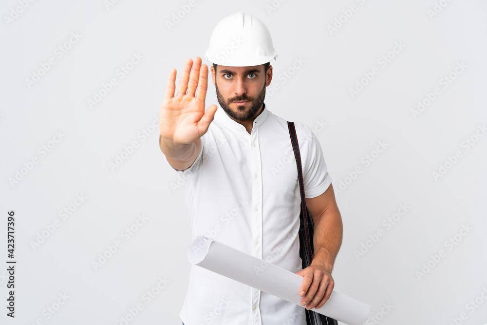 Young architect man with helmet and holding blueprints isolated on white background making stop gesture