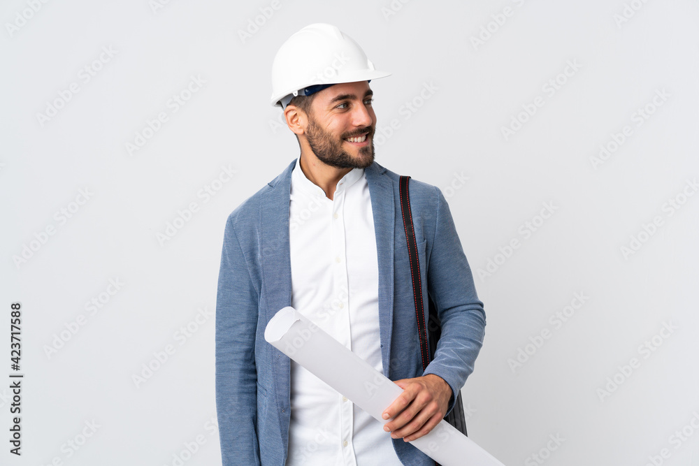 Young architect man with helmet and holding blueprints isolated on white background looking to the side and smiling Young architect man with helmet and holding blueprints isolated on white background looking to the side and smiling