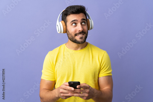 Young handsome man with beard isolated on purple background listening music with a mobile and thinking