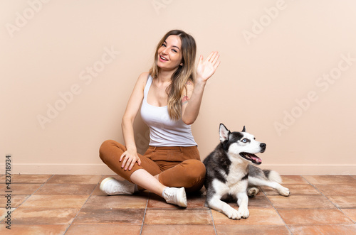 Young pretty woman with her husky dog sitting in the floor at indoors saluting with hand with happy expression