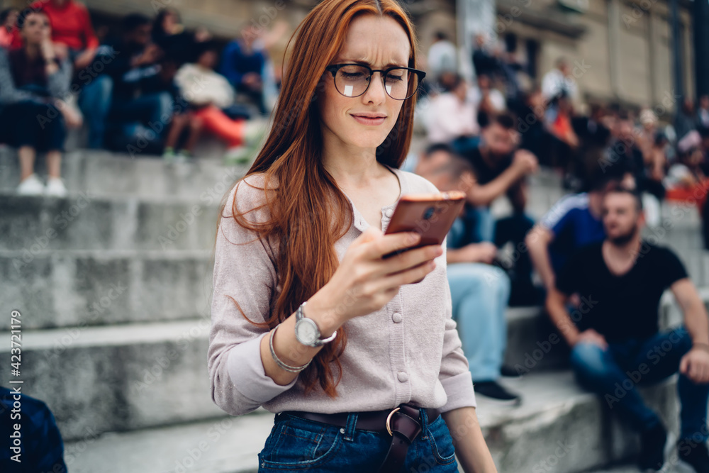 Obraz premium Puzzled female blogger in optical eyewear checking received email message with travel checklist using smartphone device with 4g internet connection, confused hipster girl making mobile booking