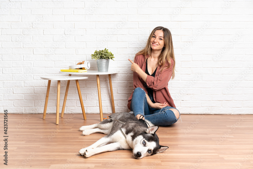Young girl with her dog sitting in the floor at indoors pointing to the side to present a product Young girl with her dog sitting in the floor at indoors pointing to the side to present a product