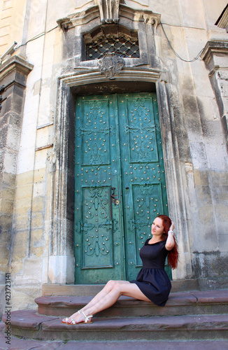 Redhead European girl sits at the ancient green door in the old European town in summer