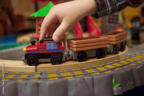 Children wooden playthings. Close up side photo of childs hand controlling toy train on the way. Little boy playing with modern wooden railway. rail road and locomotive in the play room with many toys