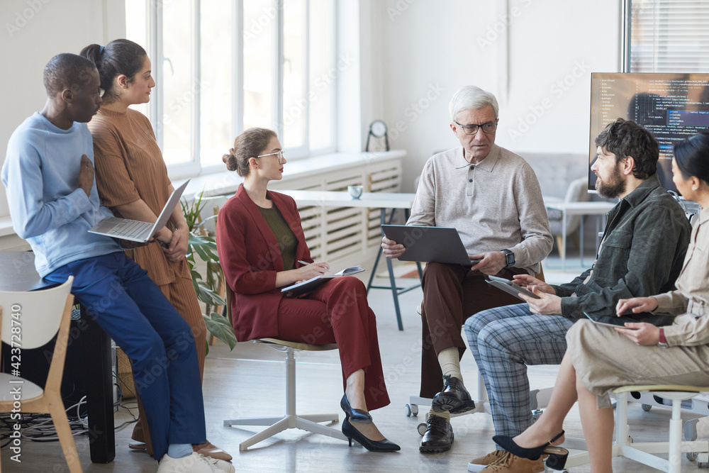 Wide angle view at contemporary diverse business team sitting in circle during strategy meeting, focus on senior businessman in center Wide angle view at contemporary diverse business team sitting in circle during strategy meeting, focus on senior businessman in center