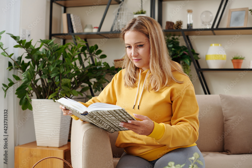Pretty young woman in yellow hoodie resting on sofa at home and reading interesting book Pretty young woman in yellow hoodie resting on sofa at home and reading interesting book