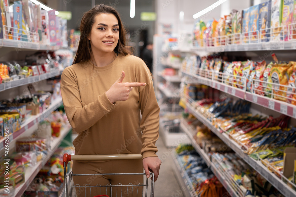 female Caucasian customer posing with shopping cart buying food in supermarket smiling looking at camera. The index finger of the hand pointed to the side. brunette choosing products in the store.