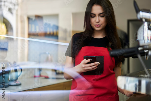 Serious 20s millennial woman in apron sending text messages on mobile phone standing at working place in coffee shop, confident female employee dialing number on smartphone at job in cafeteria