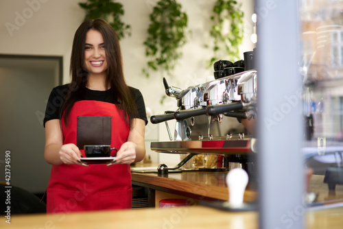 Half length portrait of happy woman waitress in apron standing near coffee machine holding cup with hot beverage  beautiful 20s prosperous female barista satisfied with job in cafeteria and bakery