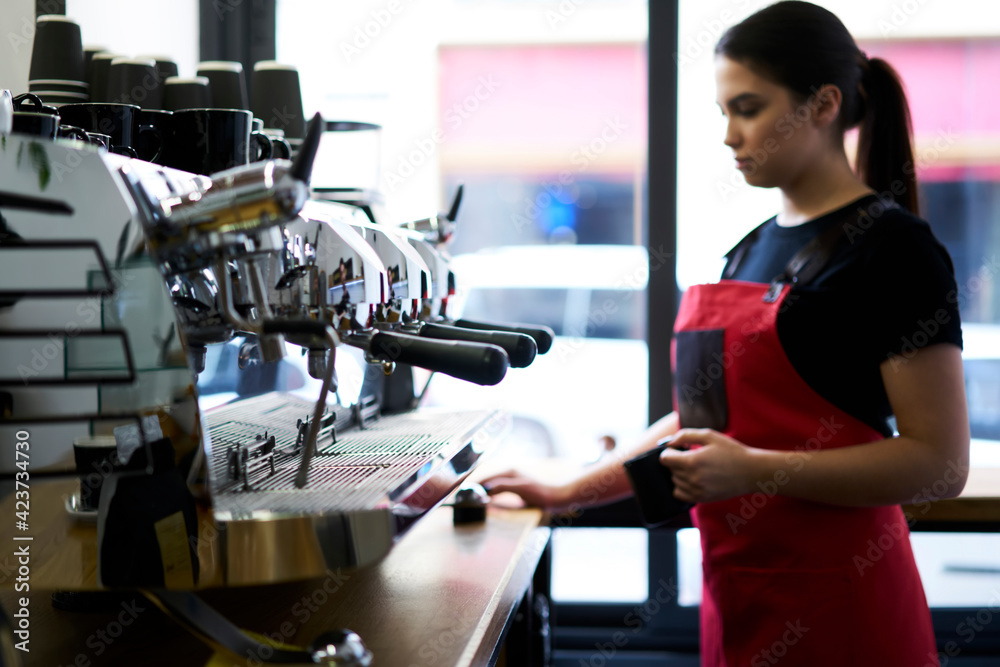 Confident female waiter in uniform using professional italian coffee machine for preparing order in cafeteria, 20s skilled woman employee making beverage on equipment holding mug at bar in cafe