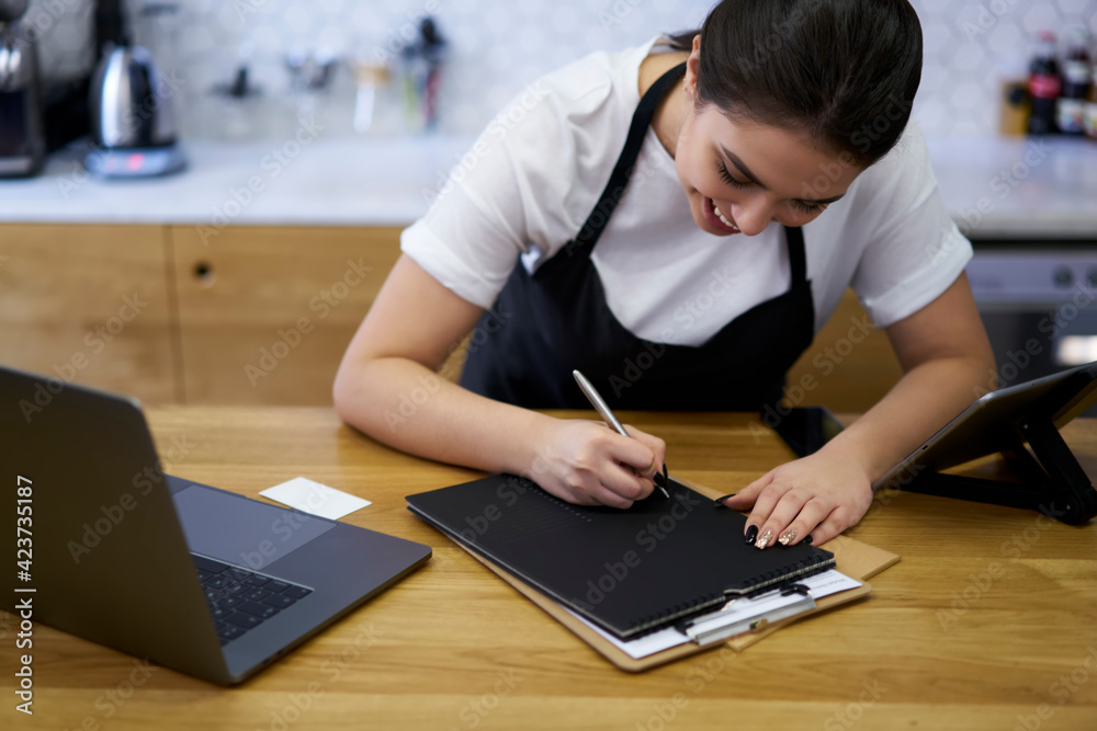 Cropped image of skilled concentrated female manager of coffee shop writing finance report and income from business near laptop computer, skilled woman barista filling Cropped image of skilled concentrated female manager of coffee shop writing finance report and income from business near laptop computer, skilled woman barista filling