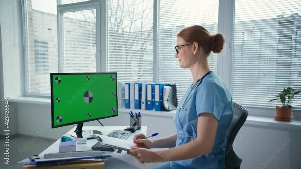 telemedicine, cute female doctor in glasses wearing blue uniform with stethoscope takes notes during remote consultation through green screen computer in health clinic