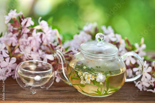 Herbal tea in a glass cup and teapot with bouquet of pink flowers on a green bokeh background. Summer tea ceremony in a garden.