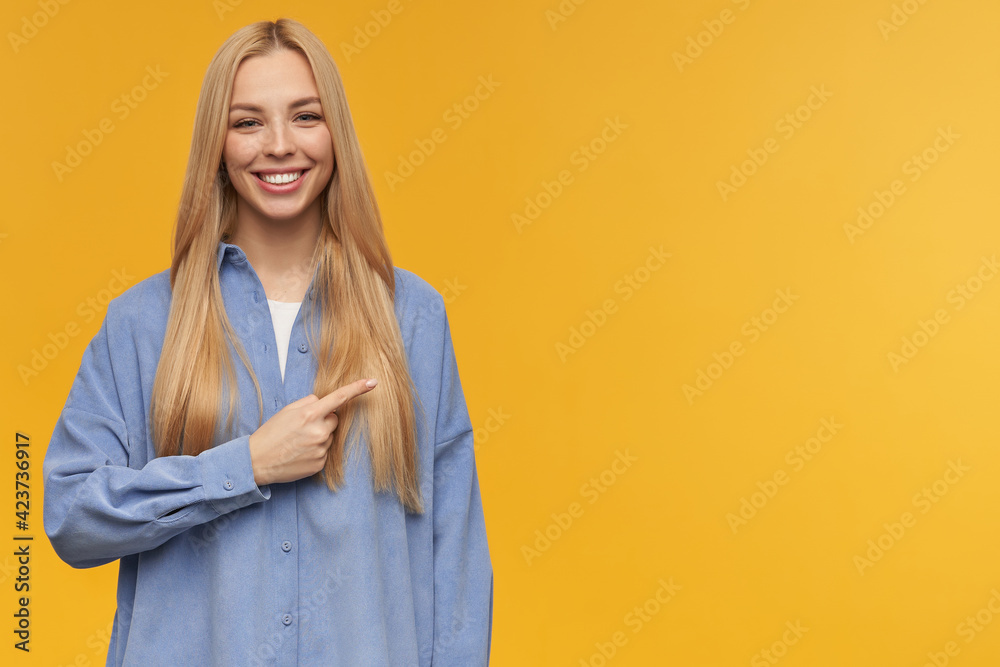 Nice looking woman, beautiful girl with long blond hair. Wearing blue shirt. People and emotion concept. Watching at the camera and pointing to the right at copy space, isolated over orange background