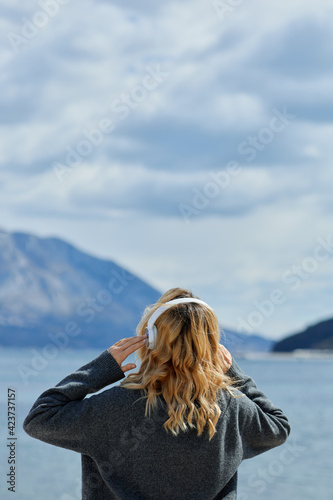 Back view of blonde woman wearing headphones listening positive music podcast from smartphone application against the sea