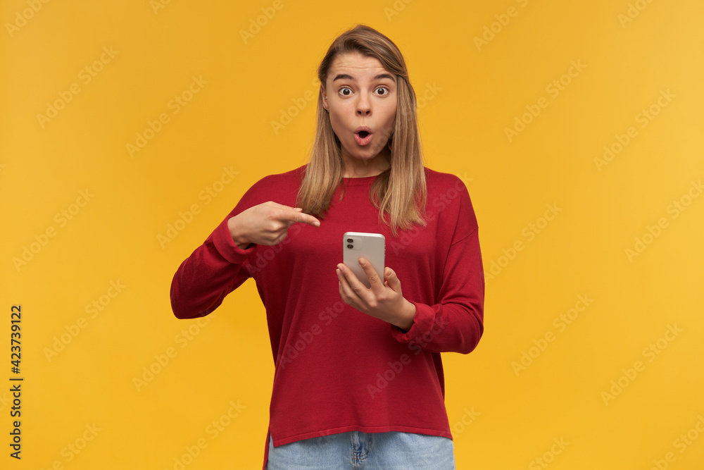 WOW! Surprised blonde young woman, points on her phone in hand, looking camera with amazement, mouth rounded, standing isolated on a yellow studio background WOW! Surprised blonde young woman, points on her phone in hand, looking camera with amazement, mouth rounded, standing isolated on a yellow studio background