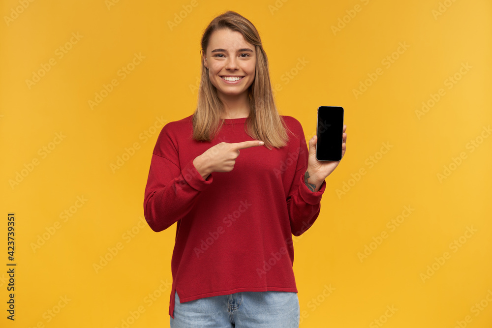 Smiling young girl keeps mobile phone in her hand, black screen turned to the camera, points on it with index finger, standing isolated on a yellow studio background Smiling young girl keeps mobile phone in her hand, black screen turned to the camera, points on it with index finger, standing isolated on a yellow studio background