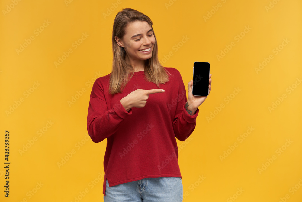 Charming young girl smiles, keeps mobile phone in her hand, black screen turned to the camera, looks on it and points with index finger, standing isolated on a yellow studio background