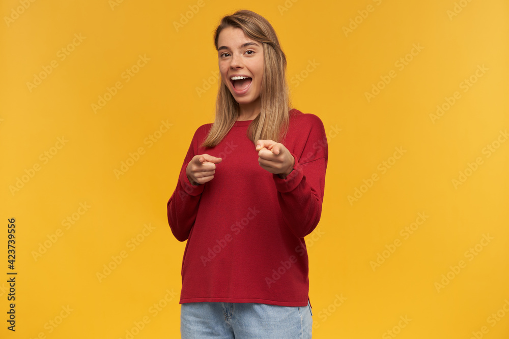 indoor shot of young female with a happy facial expression staring into camera and points forward with fingers. standing and isolated over orange background