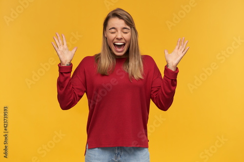Successful looking woman, rounded mouth. Winner with long blond hair. Wearing red sweater. Holding one fist up celebrating her victory. Looking camera isolated over yellow background