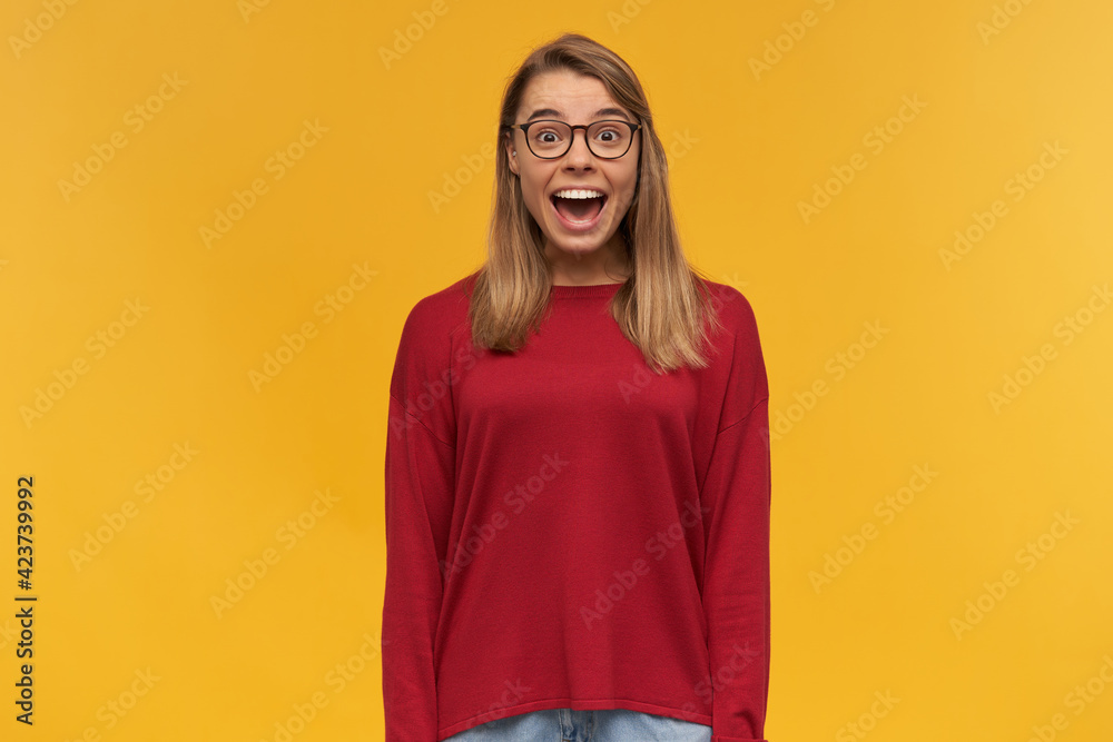 Head shot of funny happy glad blonde young girl in glasses, widely smiles, shining from happiness, completely satisfied, dressed in red sweater, glasses, on a yellow background Head shot of funny happy glad blonde young girl in glasses, widely smiles, shining from happiness, completely satisfied, dressed in red sweater, glasses, on a yellow background