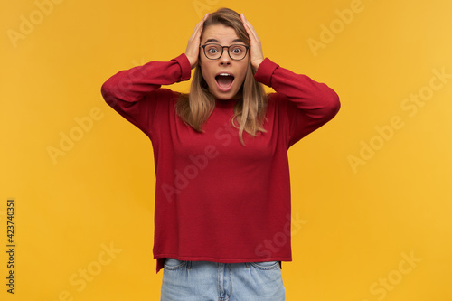 Nice looking woman, smiling girl with blond hair. Wearing red sweater and glasses. Holding fists and eyes closed. Celebrating her victory. Stand isolated over yellow background © timtimphoto