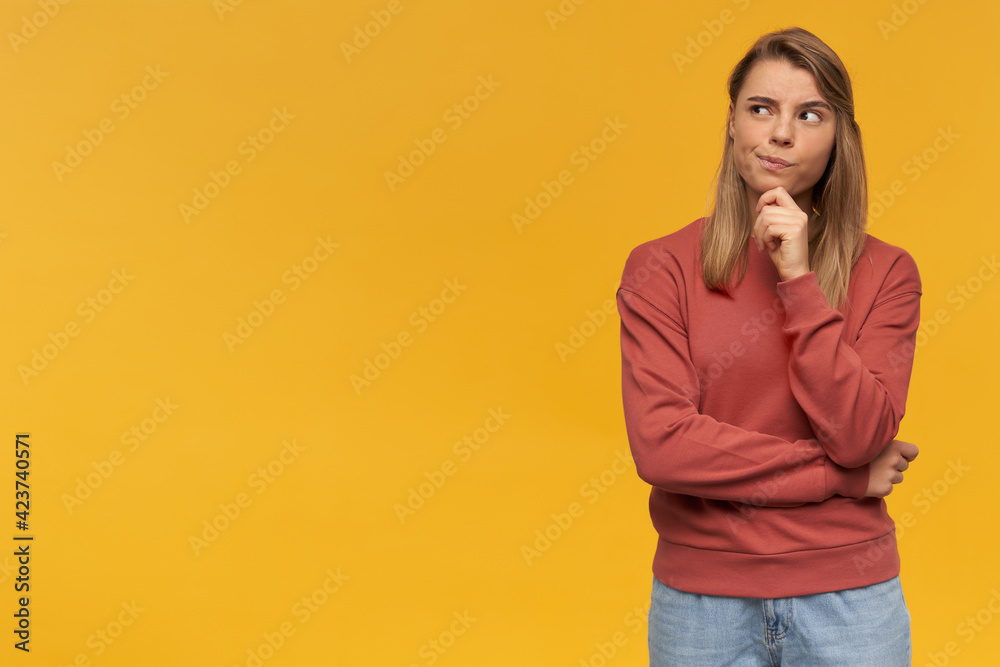 Thoughtful doubting blonde woman.Pursed lips. Looking to the upper left corner standing with hand folded on the right side isolated over yellow background. Wearing red sweater Thoughtful doubting blonde woman.Pursed lips. Looking to the upper left corner standing with hand folded on the right side isolated over yellow background. Wearing red sweater
