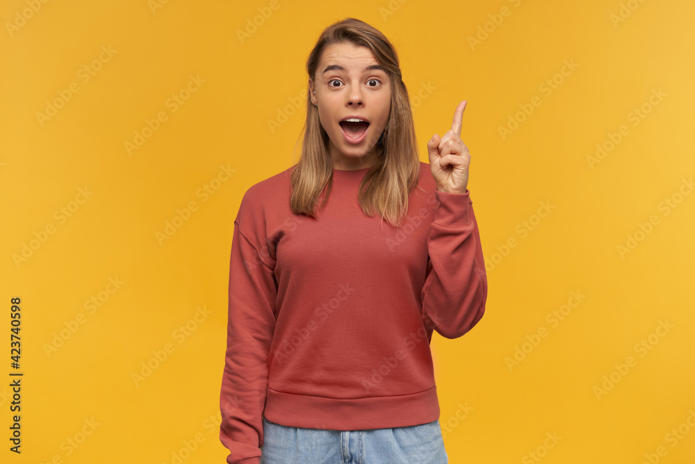 Happily looking woman, smiling girl with blond hair. Wearing red sweater and glasses. Pointing up with index finger on left hand like having a great idea.Looking camera isolated over yellow background Happily looking woman, smiling girl with blond hair. Wearing red sweater and glasses. Pointing up with index finger on left hand like having a great idea.Looking camera isolated over yellow background