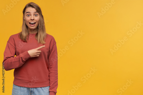 indoor shot of happy amazed young student with perfect teeth, smiles broadly, starring into camera and indicates with a finger into blank copyspace posing over orange background © timtimphoto